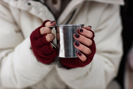 Focus on hands of young stylish woman in white winter coat and crimson mittens holding metallic mug with hot teaの写真素材