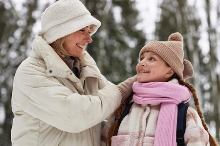 Young cheerful woman in winterwear looking at her adorable pre-teen daughter in warm cap, scarf and...