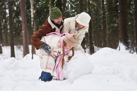 Young cheerful man in winterwear embracing his laughing wife and daughter rolling big snowball while making snowman in forestの写真素材