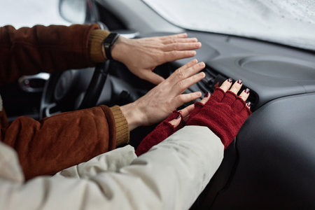 Young couple in winterwear holding hands by warm air blowing from car stove while sitting on front...