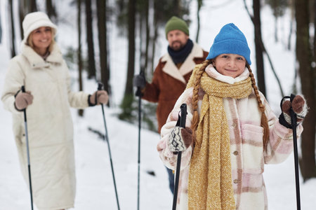 Smiling pre-teen girl with trekking sticks looking at camera while standing against her parents in park or forest on winter weekendの写真素材