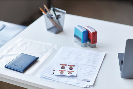 Focus on paper documents, passport and photos of applicant for visa request lying on desk with stamps, pencils and laptopの写真素材