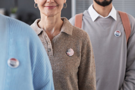 Mature smiling female voter with USA flag badge on chest standing among other people taking part in...
