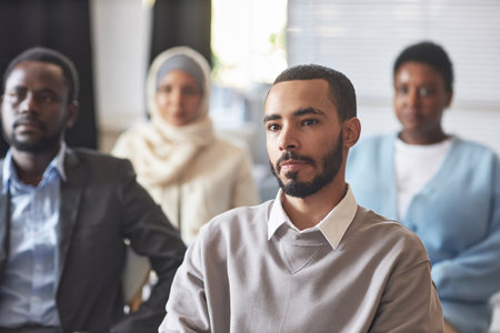 Serious multiethnic male immigrant listening to speaker at seminar or conference while sitting among...