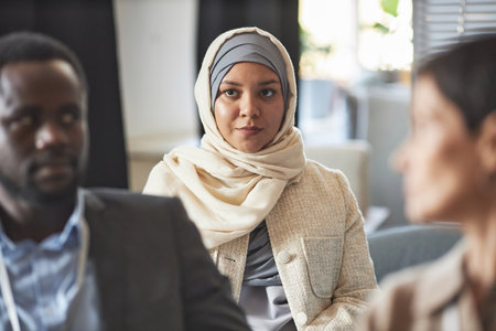Young Muslim businesswoman in hijab sitting among intercultural listeners in lecture hall during seminar or conference concerning subject of HRの写真素材