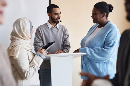 Young smiling business coach talking to intercultural female employees after seminar or training while standing by podium in lecture hallの写真素材