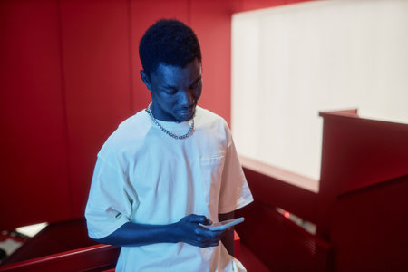 Waist up portrait of young Black man holding smartphone and scrolling social media in neon light,...