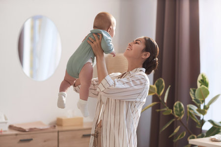 Waist up portrait of carefree young mother playing with baby son and holding him up in air, copy...