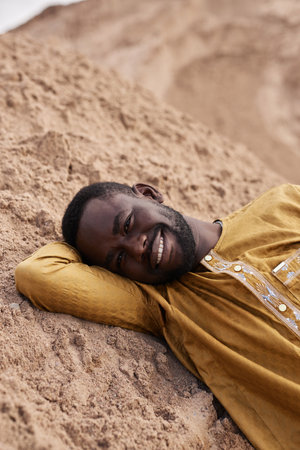 Vertical portrait of Black young man lying on sand dune in desert and smiling at cameraの写真素材