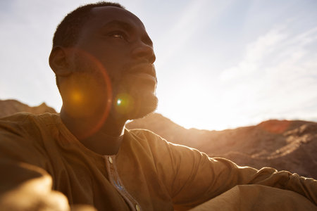 Dramatic side view portrait of Black man sitting on sand dune in desert and looking away with lens flare, copy spaceの写真素材