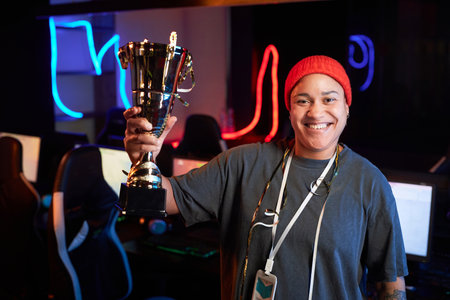 Waist up portrait of smiling Black woman holding golden cup and smiling at camera celebrating victory in eSports tournament, copy spaceの写真素材