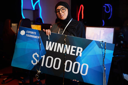 Waist up portrait of Muslim young woman holding big check and smiling at camera celebrating victory in cybersports championship, copy spaceの写真素材