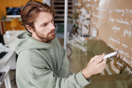 Side view at bearded young man writing data code ideas on glass in office while developing new...