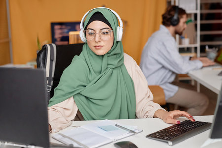 Portrait of modern Middle Eastern woman wearing headphones at workplace in office and using computerの写真素材