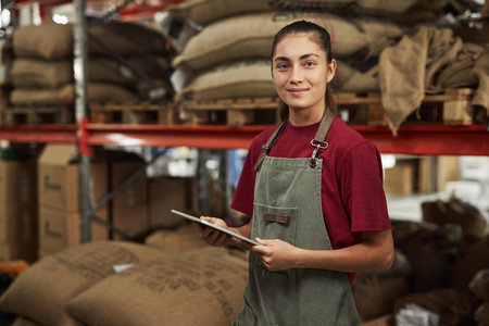 Waist up portrait of smiling young woman holding tablet and looking at camera enjoying work at warehouse in coffee roastery, copy spaceの写真素材