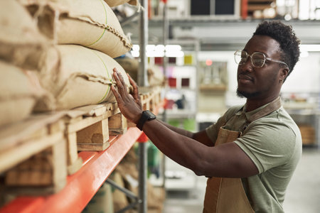 Side view portrait of Black young man inspecting burlap coffee bags on shelf in roasteryの写真素材