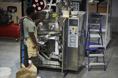 Back view of Black male worker setting up packaging machine at small coffee production factory, copy...