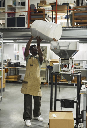 Vertical full length portrait of Black young man pouring fresh coffee beans into roasting and packaging machines at food factory, copy spaceの写真素材