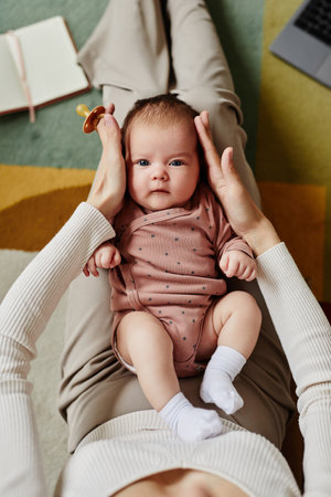 Top down view of cute baby looking at camera and lying on knees of mother holding baby head with pacifier in handの写真素材