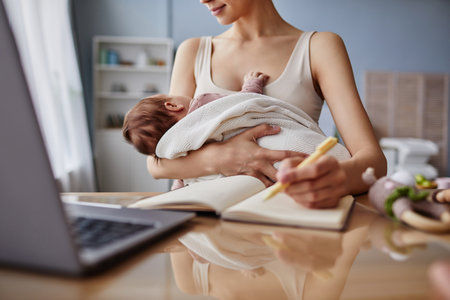 Cropped shot of unrecognizable woman with notebook sitting at desk near laptop while holding napping baby in blanket, copy spaceの写真素材