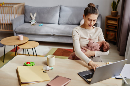 Medium shot of young Caucasian woman typing on computer while holding infant in arms in cozy room with toys, copy spaceの写真素材