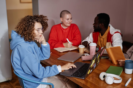 Diverse group of young people studying together at table in college dorm., focus on curly haired young woman using laptop, copy spaceの写真素材