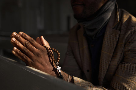 Close up of unrecognizable Black man praying in church with rosary beads in sunlight rays, copy spaceの写真素材