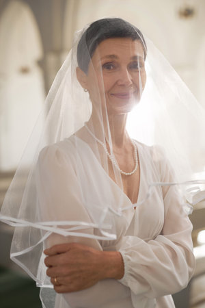 Vertical waist up portrait of beautiful adult bride wearing veil in church and smiling at cameraの写真素材