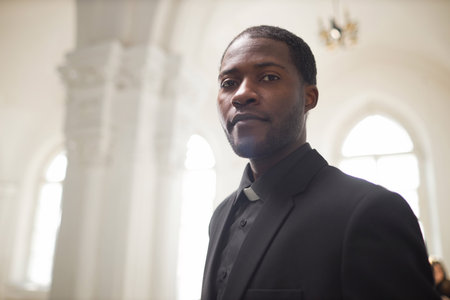 Portrait of young Black man as religious priest looking at camera in ethereal church setting, copy spaceの写真素材