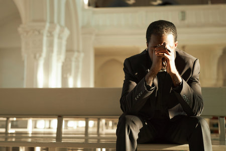 Front view at young African American priest praying in church and holding rosary beads in sunlight, copy spaceの写真素材