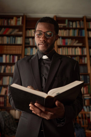 Vertical portrait of young African American man as priest reading Bible in church with books in backgroundの写真素材