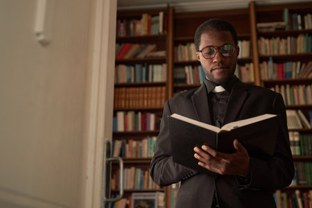 Waist up portrait of young Black priest reading Bible in office with bookcase in background, copy spaceの写真素材
