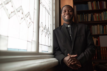 Waist up portrait of smiling African American priest looking at camera standing by window in church...