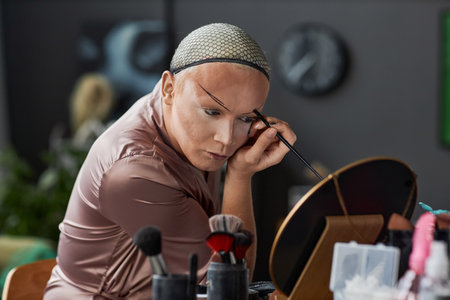 Portrait of drag queen performer doing stage makeup in dressing room and drawing on eyebrows, copy...