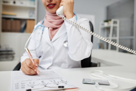 Cropped shot with selective focus on hand of female doctor using landline phone while communicating with patient remotely and making notes in clinicの写真素材