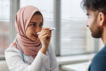 Medium close up of woman medical practitioner wearing hijab checking eyes of male patient with medical penlightの写真素材