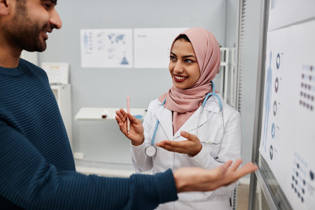 Medium of smiling Muslim young woman doctor in pink hijab talking to adult male patient showing pain intensity on medical poster in clinic officeの写真素材