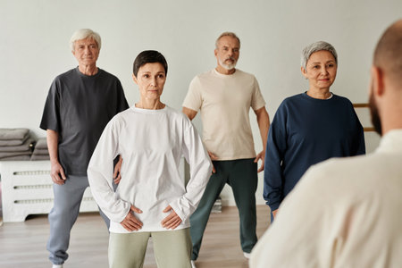 Medium long shot of senior male and female participants of fitness class looking at unrecognizable male trainer giving instructionsの写真素材