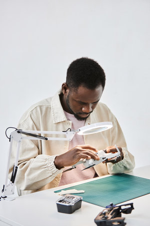 Vertical portrait of African American man inspecting drone with magnifying glass in tech repair...