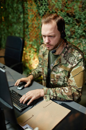Young serious male military officer in headset keeping hand on computer keyboard and looking at screen with graphic data by workplaceの写真素材