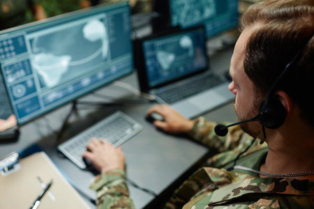 Focus on head of young military officer with headset sitting in front of computer screen with graphic data from surveillance camerasの写真素材