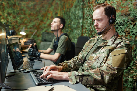 Serious young male operator in camouflage uniform typing on computer keyboard and looking at screen...