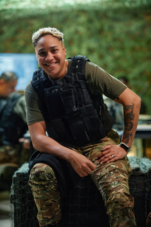 Smiling multiethnic female officer in body armor sitting on wooden chest in front of camera in surveillance room and looking at youの写真素材