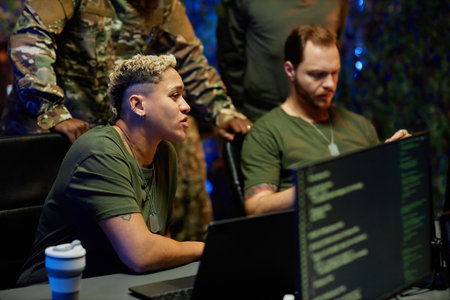 Young multiethnic female military officer sitting by workplace among male colleagues and looking at...