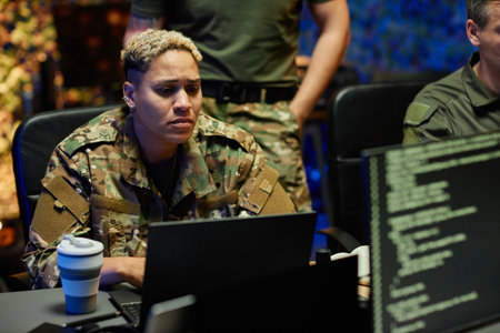 Young multiethnic female military officer in camouflage uniform looking through data on laptop screen while working among male colleaguesの写真素材