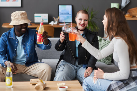 Medium full shot of three cheerful diverse young people toasting with soda while sitting on couch at...