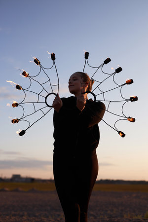 Vertical portrait of young woman holding fire fans as wings and posing against sunset sky outdoors
