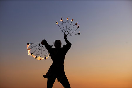 Black outline of female fire show performer dancing with flaming fans against sunset sky, copy space
