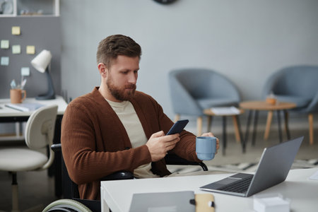Portrait of bearded young man in wheelchair working at desk in office and enjoying cup of coffee scrolling social media during break, copy spaceの写真素材