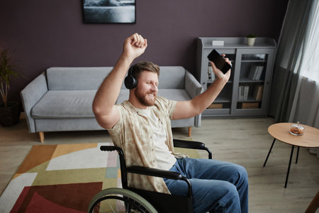 High angle portrait of carefree young man with disability dancing in wheelchair at home and listening to music, copy spaceの写真素材